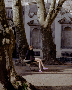 A model sitting in a piazza wearing Yuni Buffa Roma Pump shoes in Peony Pink. Comfortable artisanal crafted designer high-heels made  in Italy with Italian Lamb Nappa leather