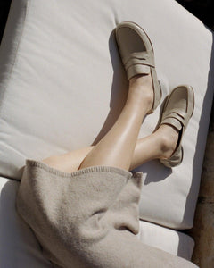 A model on a couch wearing Yuni Buffa Fez Penny Loafer shoes in Ecru beige.  Artisanal crafted women designer loafer shoe made in Italy with Italian Lamb Nappa leather.