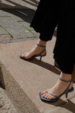 Load image into Gallery viewer, A model on the steps wearing Yuni Buffa Castrise strappy sandal Heel in Bermuda blue color handmade in Italy with Italian Lamb Nappa leather and silver logo buckle in Tuscany, Italy