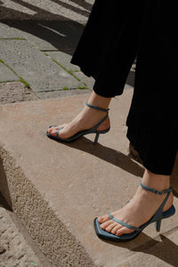 A model on the steps wearing Yuni Buffa Castrise strappy sandal Heel in Bermuda blue color handmade in Italy with Italian Lamb Nappa leather and silver logo buckle in Tuscany, Italy