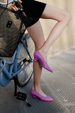 Load image into Gallery viewer, A model leaning on a bicycle wearing Yuni Buffa Roma Pump shoes in Peony Pink. Handcrafted comfortable designer high-heels made  in Italy with Italian Lamb Nappa leather