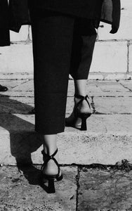 A model on the steps wearing womens designer shoe Yuni Buffa Castrise strappy sandal high heel in Black color handmade in Italy with Italian Lamb Nappa leather and silver logo buckle in Tuscany, Italy