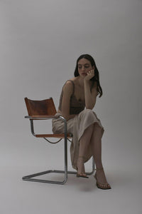 A model on leather chair wearing designer shoes Yuni Buffa Castrise strappy sandal high Heel in Sahara Beige color handmade in Italy with Italian Lamb Nappa leather and silver logo buckle