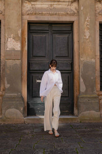 A model dressed in white walking away from a green door in a sunny tuscan town wearing Yuni Buffa Lucca Sardinian cork wedge sandal shoe in Ecru white color with tan vachetta leather insole and crepe rubber outsole made in Italy with Italian Lamb Nappa leather