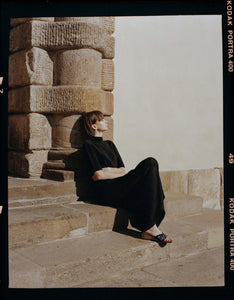 A model dressed in black sitting on  sunny Tuscan steps wearing Yuni Buffa Lucca Sardinian cork wedge sandal shoe in Navy blue color with tan vachetta leather insole and crepe rubber outsole made in Italy with Italian Lamb Nappa leather